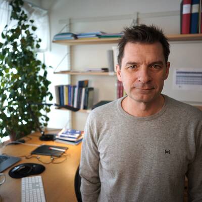 En man i grå stickad tröja tittar rakt in i kameran ståendes i sitt kontor på Umeå universitet