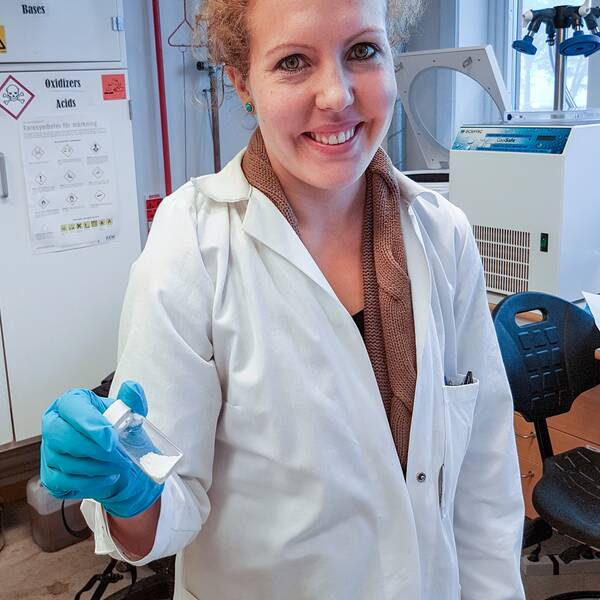 Porträtt på Sara Frykstrand doktor teknisk fysik. Hon håller en bruk med Upsalite i handen. Har en vit labbrock och blå vinylhandskat på sig. Står i labbmiljö.