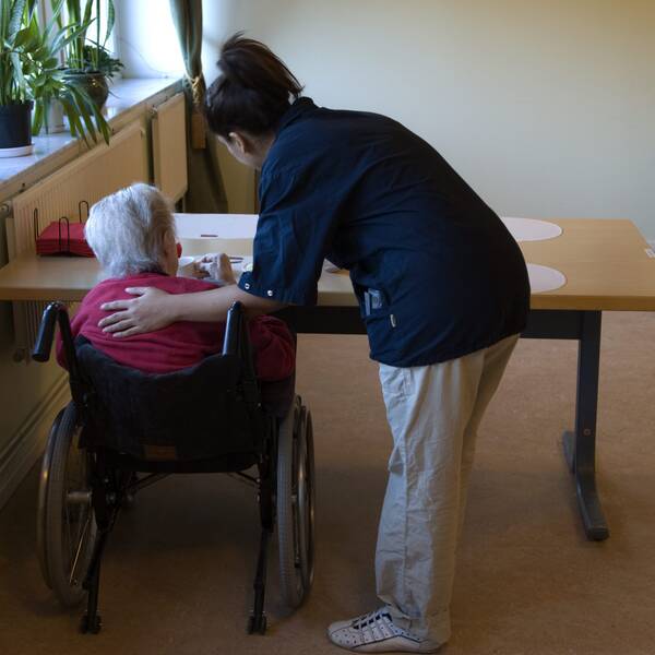 Arkivbild med äldre person i rullstol och sköterska ståendes bredvid, båda med ryggen mot kameran
