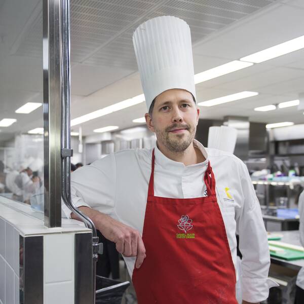Daniel Roos står i ett kök med massa kockar som arbetar bakom. Han har på sig vit rockmössa, vit kockskjorta och rött förkläde.