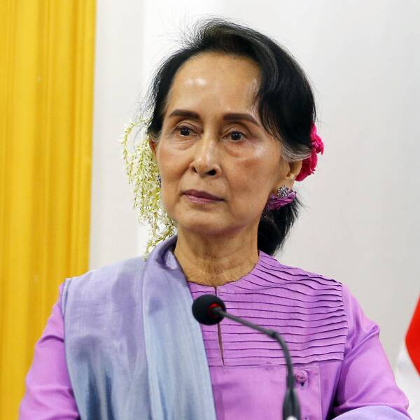 Aung San Suu Kyi står i ett talarpodium med blommor i håret.
