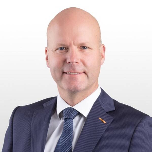Skallig man i mörkblå, kritstrecksrandig kostym. Ljus skjorta och mönstrad blå slips.