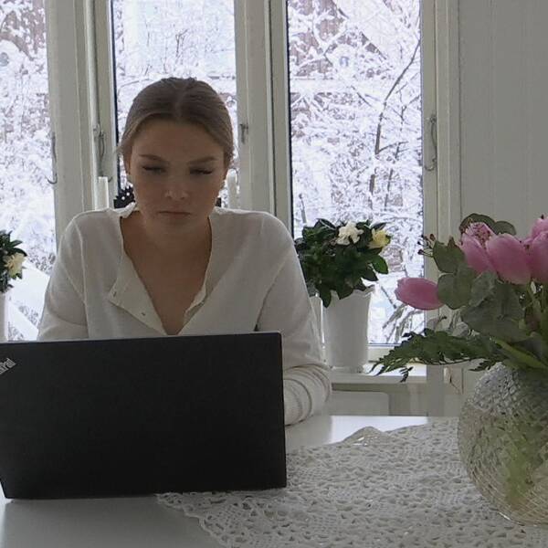 En ung kvinna sitter framför en dator vid ett köksbord.