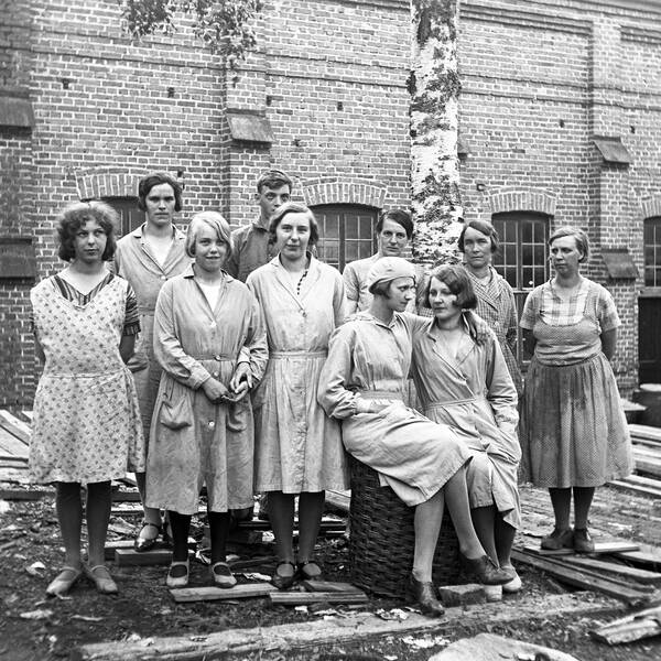 gammalt svartvitt foto av en grupp kvinnor som poserar för kameran, jord och plankor på marken, tegelbyggnad i bakgrunden