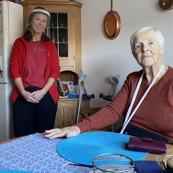 Elisabeth sitter vid sitt köksbord. I bakgrunden står Johanna Karlsson i röd hemtjänstfleece och ansiktsvisir. Elisabeth ser allvarlig ut, Johanna ser glad ut. 