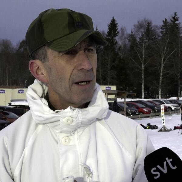 en medelålders man i vit militärjacka och grön militärkeps intervjuas framför skidspår och parkering