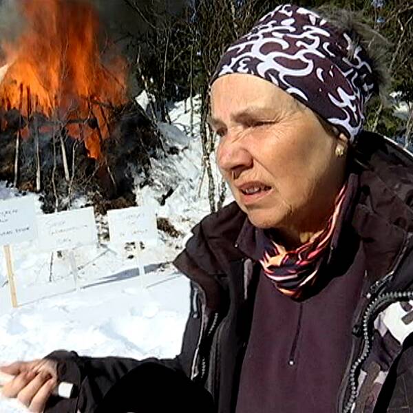 En äldre kvinna står framför en kåta som brinner