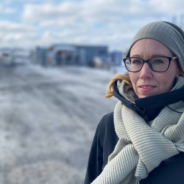 En kvinna i glasögon, halsduk och mössa, framför den nya batterifabriken i Skellefteå