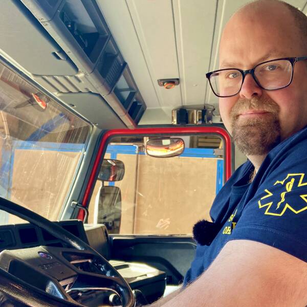 en man med glasögon klädd i tshirt med ambulanssjukvårds-märke på ärmen sitter i hytten på ett blåljusfordon, tittar mot kameran