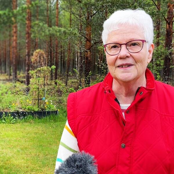 En äldre kvinna står i bild. Skogen är i bakgrunden. Hon har vitt hår och bär en röd väst.