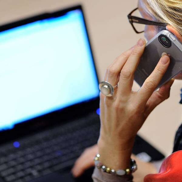 En kvinna sitter framför en dator och pratar i en smartphone.