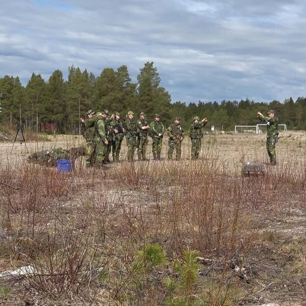 en grupp människor i gröna kamouflagekläder står på en öppen yta, skog i bakgrunden.