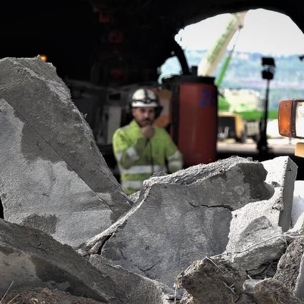 biter av riven betongstruktur, i bakgrunden en person i arbetskläder och hjälm framför en tunnelöppning