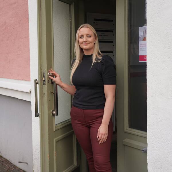 Kvinna står och håller upp en dörr, hon har blont hår, svart t-tröja och vinröda byxor. Dörren är grön och har avlånga glasrutor, huset är rosa.