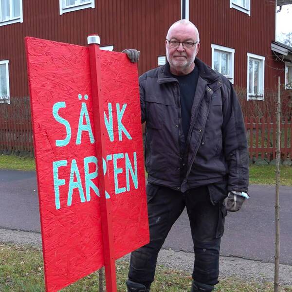 Bild på Olle Eriksson som ser bestämd ut när han står bredvid vägen och håller handen på en stor röd skylt med texten ”SÄNK FARTEN” skriven i vit text med versaler.