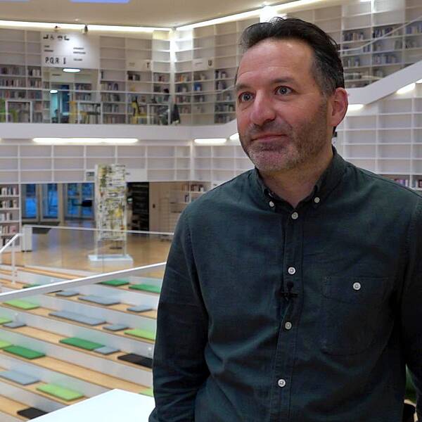 Michael Oppenheimer står i grön skjorta, högskolans bibliotek är i bakgrunden, hyllor med en massa böcker.