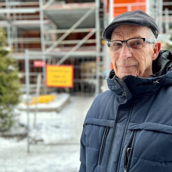 Karl-Ivar i mörkblå vinterjacka och en grå basker och glasögon. I den blurriga bakgrunden ser man ställningarna som omger huset som brandhärjades, som snart är klart. Det står även ett grönt träd till vänster.