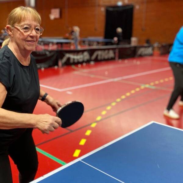 Birgitta Kronvall står framför ett blått bordtennisbord med ett pingisrack i handen. Klädd i svarta idrottskläder i en stor idrottshall med tegelväggar och rött golv.