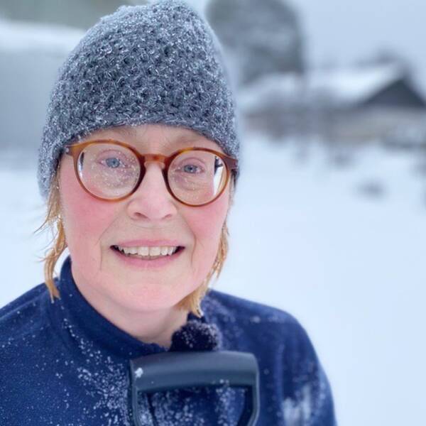 En kvinna iklädd mössa, fleecetröja och snödränkta glasögon. I nederkant anas ett spadhandtag.