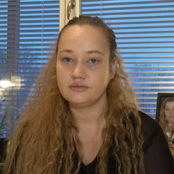 Guna Grina sitter framför ett fönster med neddragna persienner, bakom henne är en bild på hennes döda mamma.