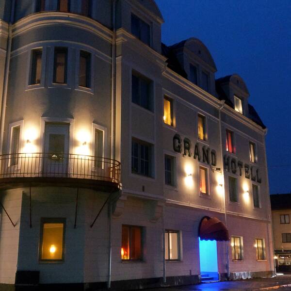 Grand hotell, i sin vita fasad, upplyst av flera vägglampor på bild tagen i kvällsmörker.