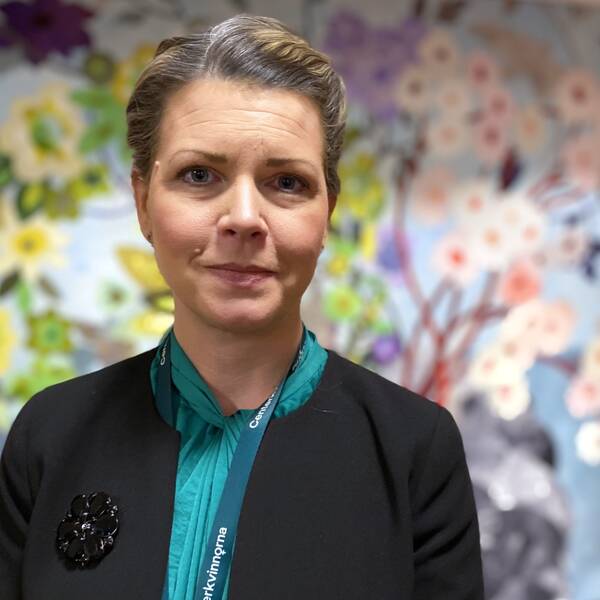 Regionrådet Sofia Jarl representerar Centerpartiet och står i entrén på regionkontoret i Falun och berättar vilken lösning hon tror på för att lösa personalkrisen inom ambulansen. Bakom henne finns en vägbonad fullt med vävda blommor.