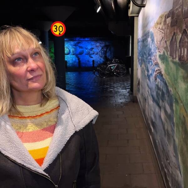 Mona Nylin, långt blont hår, mörk jacka och färgglad tröja, tittar upp på en väggmålning i garaget.