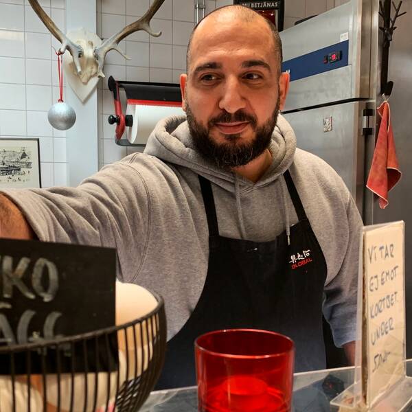 Bild på man framför restaurangkök med förklädde och gråtröja. Mannen håller i en skylt där det står eko ägg. Mannen heter  Josef Emmanuel och är en av butiksbiträdena i Saluhallen i Göteborg. Han pratar i klippet om hur inflationen påverkar