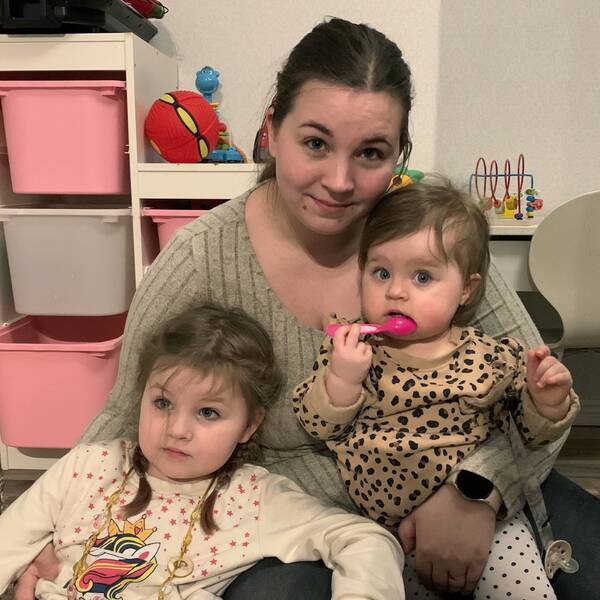 Emelie sitter på golvet i barnens lekrum i famnen håller hon döttrarna Livia, 11 månder och Ellen, 5 år. Livia har brunt kort hår och är klädde i en leopardfärgad topp. Ellen har brunt hår i flätor och har på sig en långärmad tröja med en enhörning på. I rummet och på golvet syns olika leksaker.