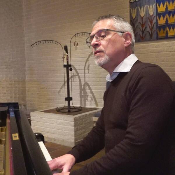 Präst sitter vid ett blanksvart piano i en kyrka och spelar samtidigt som han sjunger.