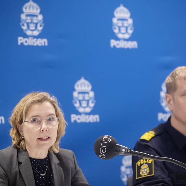 Karin Everitt har axellångt ljust hår och glasögon. Hon är iklädd svart tröja med vita prickar, grå kavaj och tittar in i kameran. Till höger om henne sitter lokalpolisområdeschefen Josef Wiklund i polisuniform. Bakom dem är en stor blå vepa med polisens logotyp i vitt.