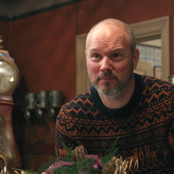 Andreas Graveleij tittar upp bakom ett stort blomsterarrangemang för att titta på det. Han står i ett rum med röda väggar och gulddetaljer.