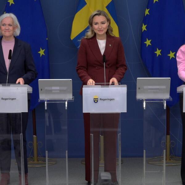 Tre kvinnor står uppradade bakom talarstolar där det står Regeringen. Bakom dem syns Sveriges och EU:s flaggor.