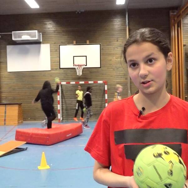 12 årig flicka i röd t-shirt som håller i en grön handboll. Bakom ser man klasskompisar som spelar handboll.