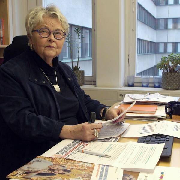 Eva Eriksson som är förbundsordförande i SPF seniorerna sitter vid sitt skrivbord. Hon har kort ljust hår, glasögon och är klädd i mörka kläder. Bakom henne finns en hylla full med böcker i olika färger och skrivbordet är fullt med olika dokument.