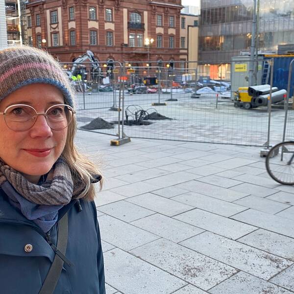 Kvinna framför byggarbetsplats på torg, en cyklist åker förbi i bakgrunden.