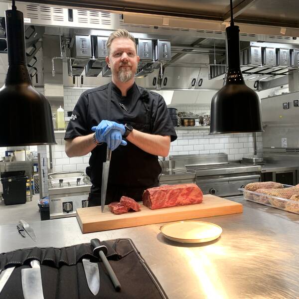 Man, Anders Törnkvist, skägg, blont hår, svart skjorta, bakom restaurangdisk. I bakgrunden kök medmaskiner och vitt kakel på väggarna.