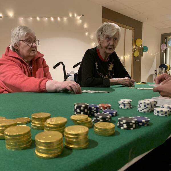 Två pensionärer på ett äldreboende sitter och spelar kort med spelmarker på det gröna spelbordet framför sig.