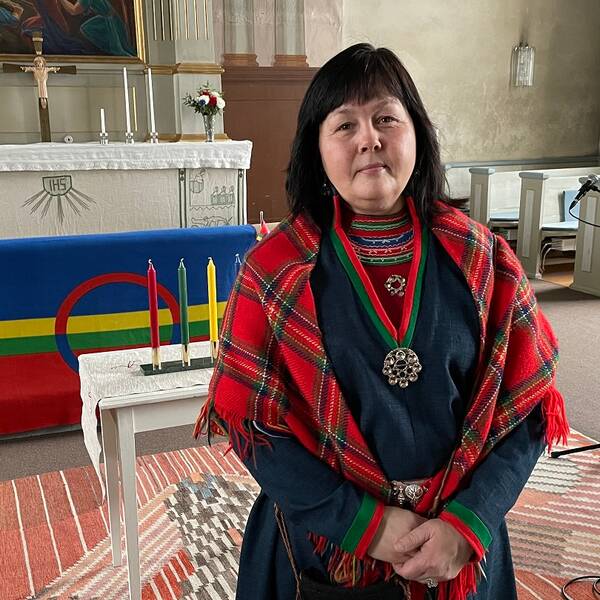 Aanna Johansson Larsson, är samisk samordnare i Strömsunds kommun. Hör henne berätta varför det är så viktigt att fira Samefolkens dag. På bilden ser man Aanna Johansson Larsson stå i en kyrka. Hon är klädd i en sydsamisk kolt som är mörkblå med röda och gröna revärer. Över axlarna har hon en rödrutig sjal. Hon har mörkt långt hår.