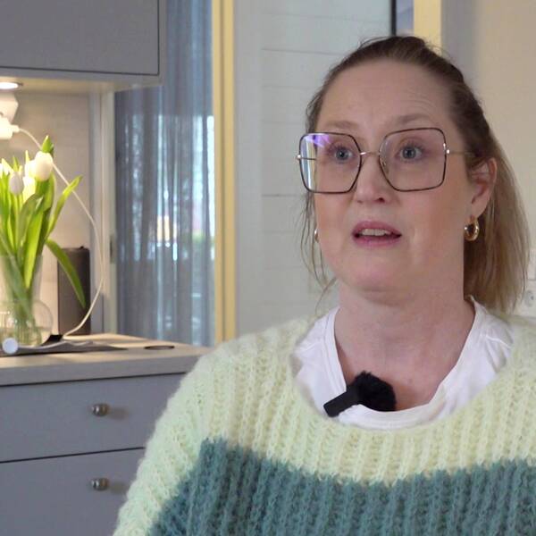 En kvinna i stickad tröja och stora glasögon sitter i en köksliknande miljö.