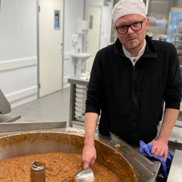 Bild på en man i 50-årsålden med kockmössa och svarta glasögon. Han står framför en stor gryta med köttfärssås som han rör om i. I bakgrunden skymtar ett storkök.