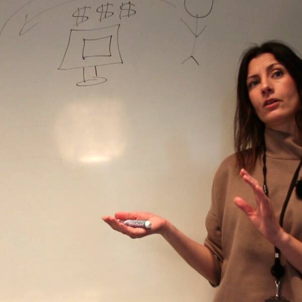 Kvinna framför whiteboardtavla med streckgubbar, en dator och dollartecken.
