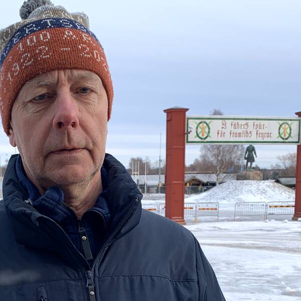 Vasaloppets sportschef Tommyhöglund står till höger i bild med blå jacka och roströd ocvh grå stickad mössa. Vasaloppsmålet syns bakom, höger i bild, där står det: I fäders spår – för framtids segrar.