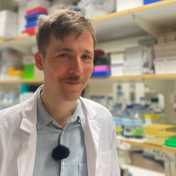 På bilden syns en man med blå skjorta och vit rock. Mannen heter Daniel Engelbertsen och han är forskare vid Lunds universitet. På bilden befinner han sig inne i ett labb.