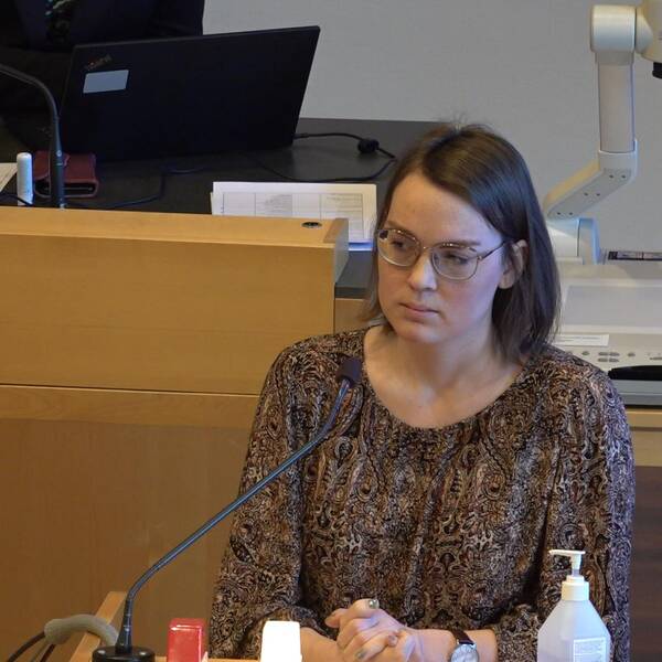 En kvinna i brun page och brun paisleymönsterad blus står bakom en mikrofon och ser ledsen ut.