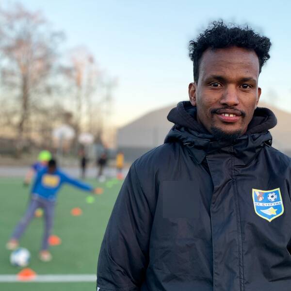 Fotbollstränaren och grundaren av fotbollsklubben BK Ett Fyra, Daud Awees, står på en fotbollsplan i klubbjackan medan det pågår en träning bakom honom.