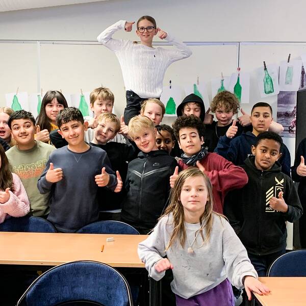 En bild på cirka 20 barn i ett klassrum, de ser glada ut och gör tummen upp. 