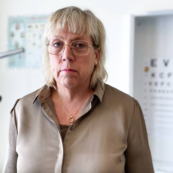 Ingmarie Kasche står i sitt arbetsrum. I bakgrunden syns en brits och en karta med bokstäver i olika storlekar som används vid synundersökningar.