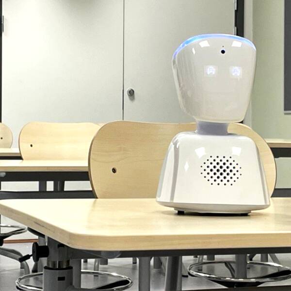 En vit oformlig liten robot står i ett klassrum med glada ögon.
