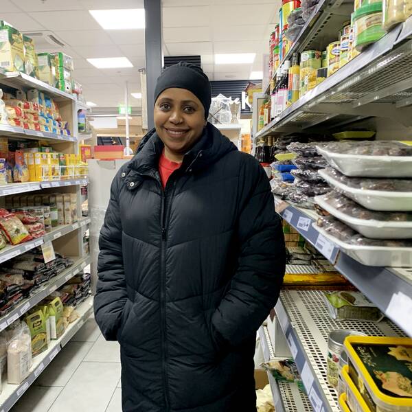 Fatime Noor, står mellan två hyllor mat i en matbutik, är mörkhyad, har svart huvudbonad och jacka, röd tröja, hon ler mot kameran.